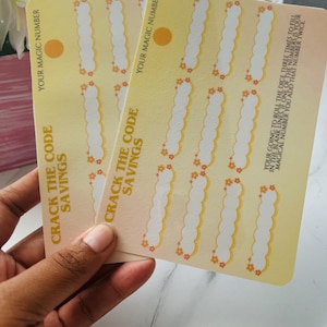 Könnte beinhalten: Zwei gelbe Karten mit dem Text "CRACK THE CODE SAVINGS" und "YOUR MAGIC NUMBER". Jede Karte hat mehrere leere Felder mit einem floralen Design. Die Karten werden in einer Hand gehalten.