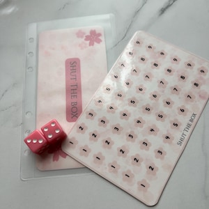 Puede incluir: Una tarjeta de juego rosa y blanca con el texto "SHUT THE BOX" y los números del 1 al 9. Dos dados rosas están sobre una tarjeta rosa y blanca con el texto "SHUT THE BOX".
