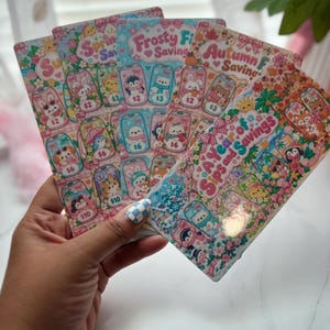 May include: A set of four rectangular paper savings trackers featuring colorful illustrations of soda cans with animal characters. The cards are titled Spring Savings, Frosty Fun Savings, Autumn Savings, and A Year of Sips and Savings, with various dollar amounts printed on each individual can graphic.