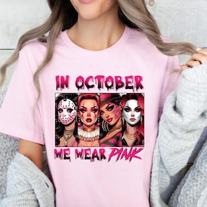 K&ouml;nnte beinhalten: Pinkes T-Shirt mit einem Grafikdruck von vier Frauen, die Halloween-Masken tragen. Der Text "In October We Wear Pink" ist auf dem Shirt aufgedruckt.
