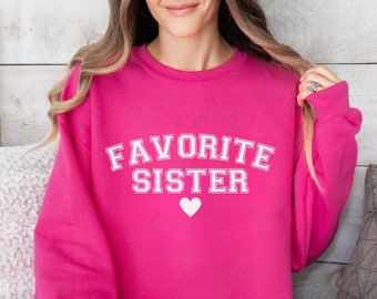 May include: A pink sweatshirt with the text "FAVORITE SISTER" in white letters and a small white heart.