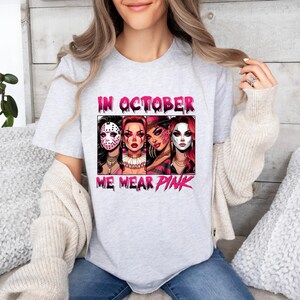 K&ouml;nnte beinhalten: Ein graues T-Shirt mit einem pinkfarbenen Grafikdesign, das vier Frauen zeigt, die Horrorfilm-Masken tragen. Der Text "In October We Wear Pink" ist auf dem Shirt aufgedruckt.
