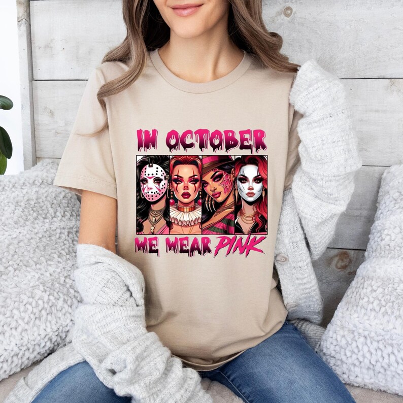 K&ouml;nnte beinhalten: Ein beiges T-Shirt mit dem Text "In October We Wear Pink" in rosa Schrift. Das Shirt zeigt eine Grafik von vier Frauen in Halloween-Kost&uuml;men mit rosa Akzenten.