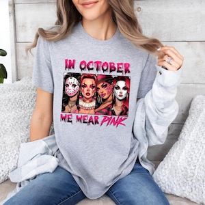 K&ouml;nnte beinhalten: Graues T-Shirt mit einer pinkfarbenen Grafik, auf der "Im Oktober tragen wir Pink" steht. Die Grafik zeigt vier Frauen in Halloween-Kost&uuml;men mit pinkfarbenen Akzenten.