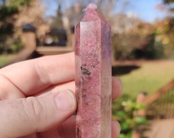 May include: A pink crystal point with a faceted design. The crystal is held in a hand and has a smooth, polished surface. The crystal is approximately 4 inches in length.