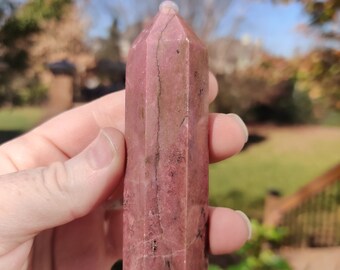 May include: A pink and brown rhodonite crystal tower. The polished gemstone has a rectangular shape with a pointed top. The crystal is held in a hand, with a blurred outdoor background.
