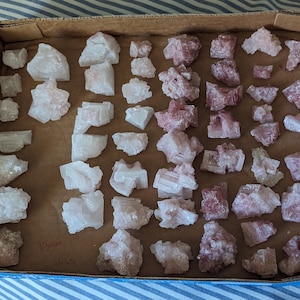 Puede incluir: Una colección de cristales de sal rosa y blanca dispuestos en una caja de cartón. Los cristales son de varios tamaños y formas, algunos son más alargados que otros. Los cristales están dispuestos en dos filas, con los cristales rosas en la fila superior y los cristales blancos en la fila inferior.