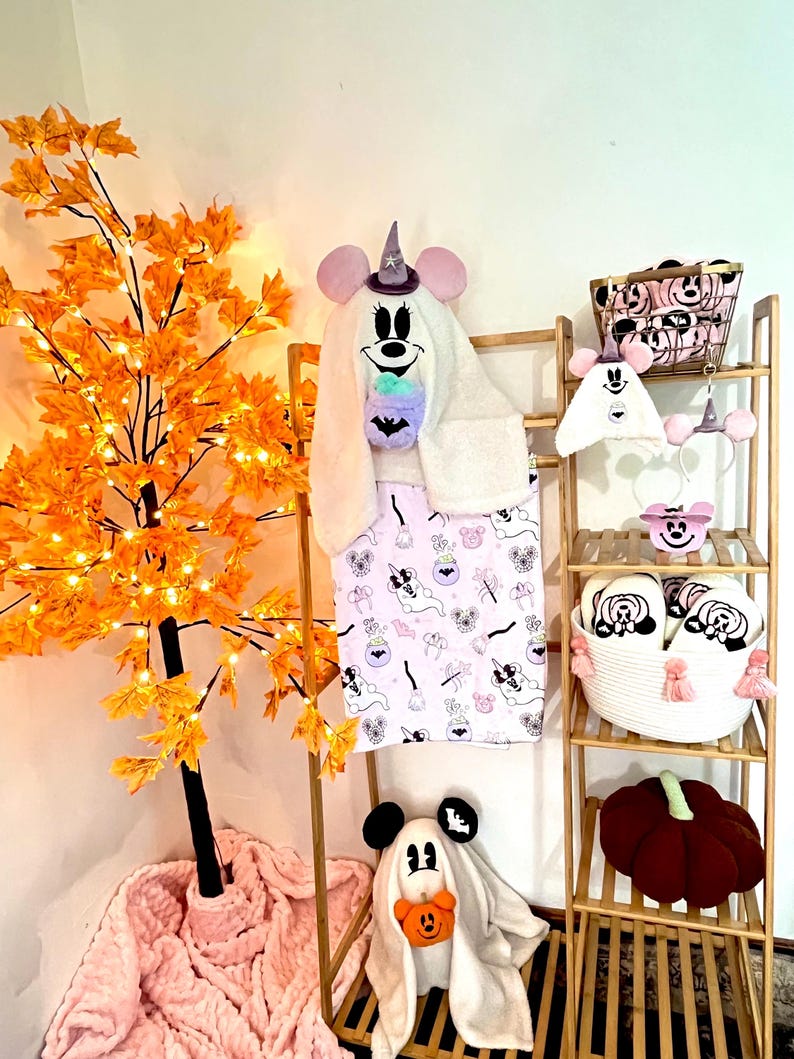 May include: Halloween-themed items, including a hooded towel, blanket, slippers, and decorative basket, all featuring a cartoon mouse design. A plush pumpkin and a decorative tree with orange leaves and lights are also visible.