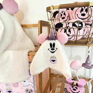 以下が含まれることがあります： 紫色の魔女の帽子と笑顔の顔が付いた、白いぬいぐるみのお化けの形をしたキーホルダー。お化けは黒い顔と紫色の大釜のデザインをしています。その他のアイテムには、ピンクのミニーマウスのヘッドバンドと、ミニーマウスの顔が付いたピンクのタオルのバスケットが含まれます。