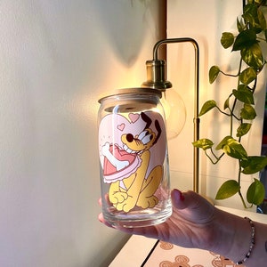 Puede incluir: Una taza de vidrio con una imagen de dibujos animados de un perro sosteniendo un hueso en forma de corazón. El perro es amarillo y el corazón es rosa. La taza está sobre una mesa con una superficie de patrón marrón y blanco.