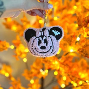May include: A pink plush keychain shaped like a cartoon character's head, featuring black ears and facial details. A white bat graphic is on one ear. The keychain is held by a gold-colored clasp.