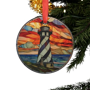May include: A round glass ornament with a stained glass design of a lighthouse against a sunset sky. The lighthouse is white with black stripes and has a red roof. The sky is orange, yellow, and blue. The ornament is perfect for a nautical themed Christmas tree.