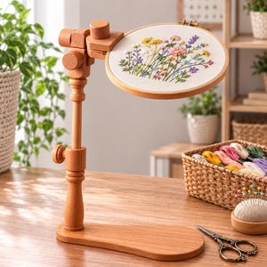 May include: A wooden embroidery hoop stand with an adjustable arm and base, holding a circular hoop containing white fabric with a colorful floral embroidery design. Beside it are a basket of embroidery floss, a pincushion, and scissors.