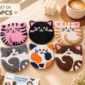 May include: Set of six cat-shaped coasters in various colors, including pink, black, and brown. Each coaster features a different cat face design, with white whiskers and a small pink nose. The coasters are made with a textured, looped fabric.