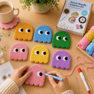 May include: A collection of colorful, handmade ghost-shaped coasters in yellow, blue, green, pink, and purple. A kit with yarn and tools is also visible. The coasters are on a wooden surface, with a cup of coffee and a plant in the background.