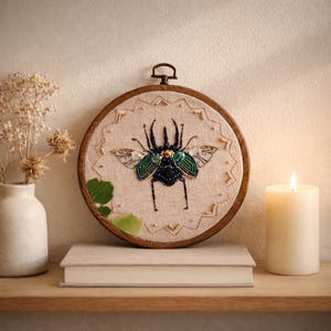 May include: A handcrafted embroidery hoop featuring a detailed beetle design. The insect is embellished with beads in shades of blue, green, and gold, set against a beige fabric. The wooden hoop is displayed on a shelf with a candle and a vase of dried flowers.