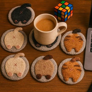 May include: A collection of six circular tufted coasters featuring various cat rear designs in black, cream, orange, and brown colors. One coaster supports a beige ceramic mug filled with coffee, while a colorful puzzle cube sits nearby.