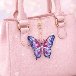 May include: A pink handbag with gold-tone hardware and a butterfly charm. The butterfly charm is embellished with blue, purple, and pink rhinestones, and hangs from a gold chain.