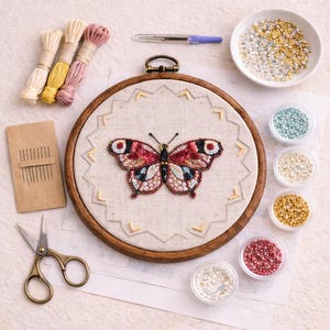 May include: A craft kit featuring a wooden embroidery hoop with a beaded butterfly design. The kit includes embroidery floss, needles, scissors, and small containers of beads in various colors. A pen is also included.