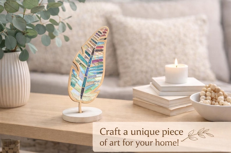 Op de afbeelding: Een decoratieve veersculptuur met een moza&iuml;ekontwerp in blauwe, groene en paarse tinten. De veer is gemonteerd op een houten voet. Een brandende kaars, boeken en een kom met kralen staan ook op de tafel. De tekst luidt: "Maak een uniek kunstwerk voor je huis!"