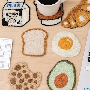 May include: A collection of whimsical, textured coasters in various food shapes, including a milk carton with a cow, a slice of bread, a fried egg, a cookie, and an avocado. A white mug filled with coffee sits on a coaster. The coasters are on a light wood surface.