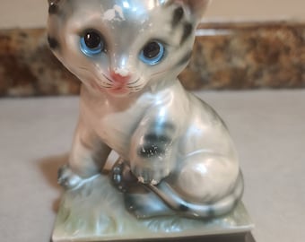 May include: A ceramic figurine of a gray and white tabby kitten with blue eyes. The kitten is sitting on a green base with a black base.