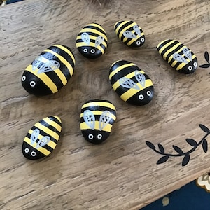 Pode incluir: Sete pedras pintadas em forma de abelha sobre uma superfície de madeira. As pedras são pintadas de preto e amarelo com asas cinzas e olhos pretos.