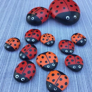 Puede incluir: Una colección de mariquitas pintadas, cada una con puntos negros y ojos blancos, sobre un fondo gris texturizado. Las mariquitas están pintadas sobre piedras lisas y redondas.