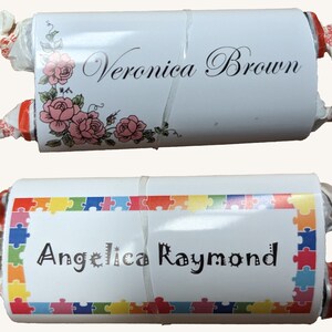May include: Two white, cylindrical candy wrappers with text. One wrapper reads "Veronica Brown" with pink roses. The other wrapper reads "Angelica Raymond" with a colorful puzzle border. Both are wrapped with white and red twist ends.