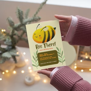 May include: A person holds a cream-coloured invitation card featuring a cartoon bee and the words "Bee There!". The card details Amy's birthday party, including the date, time, and location. Green leaf accents are used to frame the text on the card.
