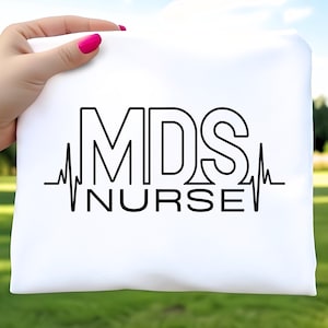 May include: A white t-shirt with the text "MDS NURSE" in black, with a heartbeat line graphic underneath. The shirt is held up against a blurred green background.