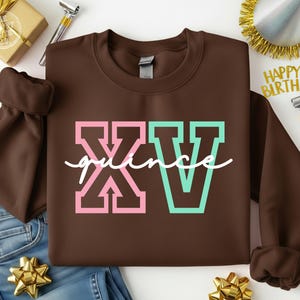 Puede incluir: Una sudadera marr&oacute;n con el texto "XV quince" en letras rosas y turquesas. La sudadera se muestra sobre una superficie blanca con una caja de regalo dorada, adornos de fiesta y vaqueros azules.
