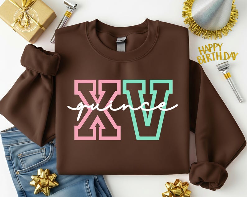 Puede incluir: Una sudadera marr&oacute;n con el texto "XV quince" en letras rosas y turquesas. La sudadera se muestra sobre una superficie blanca con una caja de regalo dorada, adornos de fiesta y vaqueros azules.