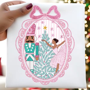 May include: White fabric with a pink glittery bow and oval frame design. Inside the frame is a Christmas scene with a nutcracker, a ballerina, and a Christmas tree. The nutcracker is in a green and white uniform. The ballerina is wearing a pink dress.