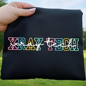 Könnte beinhalten: Schwarze Tasche mit dem Text "XRAY TECH" in bunten Blockbuchstaben. Das Wort "XRAY" ist in Rosa, Blau und Grün, während "TECH" in Grün, Blau und Gelb gehalten ist. Die Wörter sind in einer fetten, umrandeten Schrift.