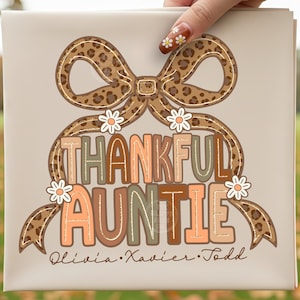 May include: A beige square canvas with the words "THANKFUL AUNTIE" in colorful, layered letters. A leopard print bow and daisy accents frame the text. The names Olivia, Xavier, and Todd are written below.