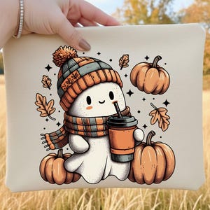 以下が含まれることがあります： ニット帽とスカーフを身につけ、コーヒーカップを持った漫画の幽霊が描かれたベージュのクラッチバッグ。カボチャと秋の葉が幽霊を囲んでいます。デザインはオレンジ、グレー、白の色で、秋にぴったりです。