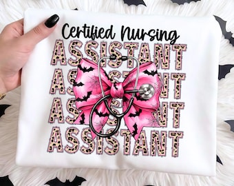 May include: A white t-shirt featuring the text Certified Nursing ASSISTANT in a repeating leopard print pattern. A large pink bow with black bat and spiderweb graphics is centred over the text, with a black medical stethoscope draped over it.