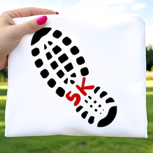 May include: White t-shirt featuring a black shoe print with the red text "5K". The design is held by a hand with pink nail polish, set against a blurred green background.