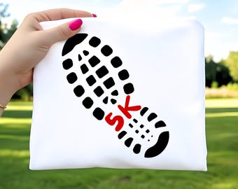 Op de afbeelding: Wit T-shirt met een zwarte schoenafdruk en de rode tekst "5K". Het ontwerp wordt vastgehouden door een hand met roze nagellak, tegen een vage groene achtergrond.