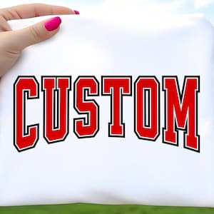 Op de afbeelding: Een wit t-shirt met het woord CUSTOM gedrukt in een vet, gebogen lettertype in varsity-stijl. De letters zijn rood met een dikke zwarte omlijning, wat zorgt voor een grafisch ontwerp met hoog contrast op de voorzijde van het kledingstuk.