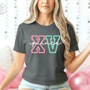 Puede incluir: Camiseta gris oscuro con la palabra "XV" en letras de bloque rosas y turquesas, y la palabra "quince" en escritura blanca. La camiseta es de cuello redondo y tiene mangas cortas. La camiseta es de estilo casual.