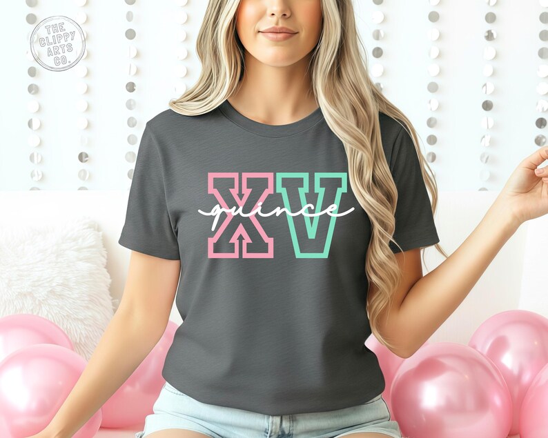 Puede incluir: Camiseta gris oscuro con la palabra "XV" en letras de bloque rosas y turquesas, y la palabra "quince" en escritura blanca. La camiseta es de cuello redondo y tiene mangas cortas. La camiseta es de estilo casual.