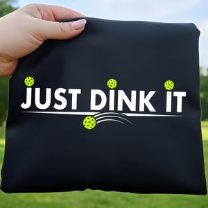 May include: Black tote bag with the text "JUST DINK IT" in white, along with yellow pickleball graphics. The bag is held up against a green grassy background.