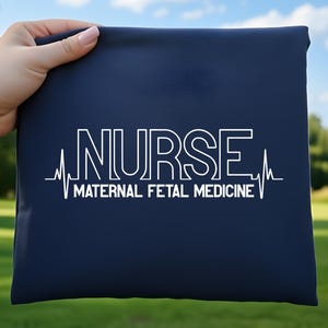 May include: A navy blue bag with white text that reads "NURSE MATERNAL FETAL MEDICINE" with a heartbeat line graphic. The bag is held up against a background of green grass and trees.