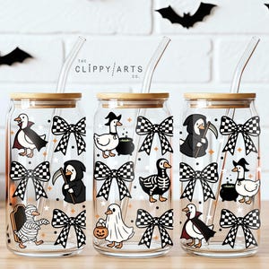 May include: Three clear glass tumblers with bamboo lids and glass straws, featuring a repeating pattern of ducks dressed in Halloween costumes, including ghosts, mummies, and skeletons, interspersed with black and white checkered bows.