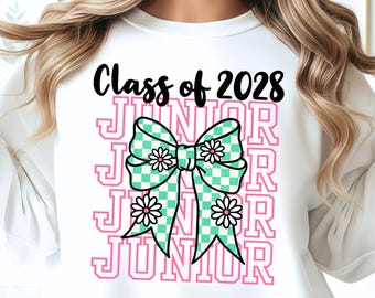 May include: A white sweatshirt featuring a graphic print with the black text Class of 2028 above a large teal and white checkered bow. The bow is decorated with white daisies and layered over the pink outlined text JUNIOR repeated four times.