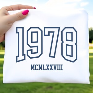 May include: A white t-shirt with the year "1978" printed in navy blue, along with the Roman numeral "MCMLXXVIII" below. The shirt is held up by a hand with pink nail polish, set against a blurred green background.