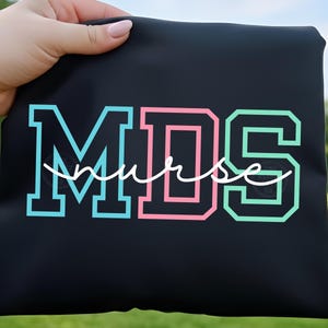 May include: Black tote bag with the text "MDS nurse" in teal, pink, and green block letters with a white cursive font. The bag is held up against a blurred green background.