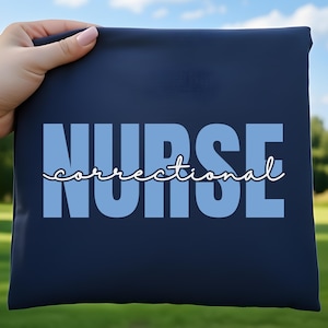 May include: A dark navy blue t-shirt featuring the word NURSE in large, light blue block letters. The word correctional is written in a white, cursive script font layered horizontally across the center of the block letters.